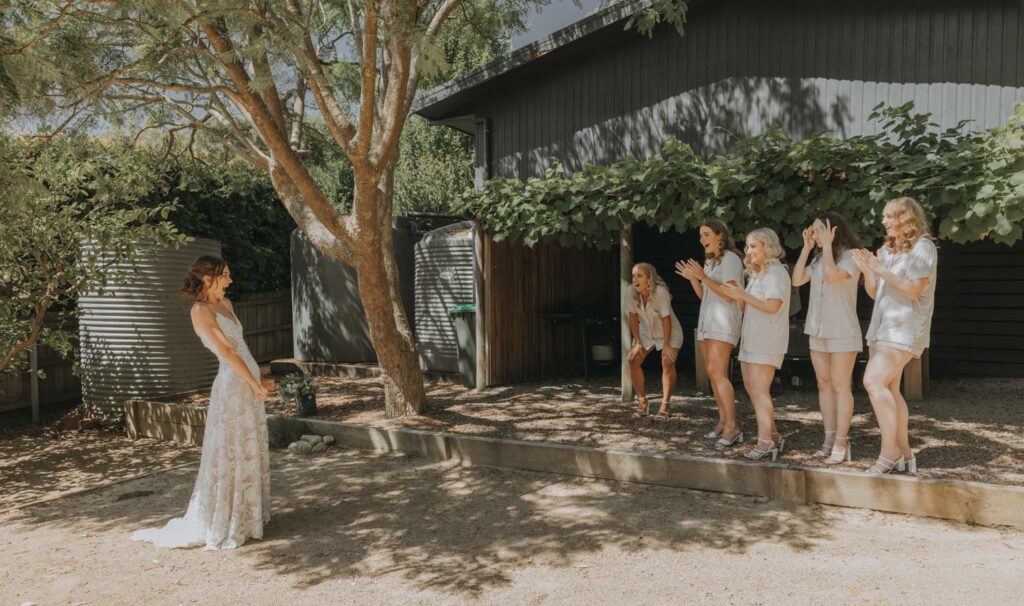 A bride in a lace wedding dress stands serenely under the shade of a tree, facing away from a group of bridesmaids dressed in white, who are applauding and expressing joy, likely captured by a wedding photographer at an outdoor ceremony