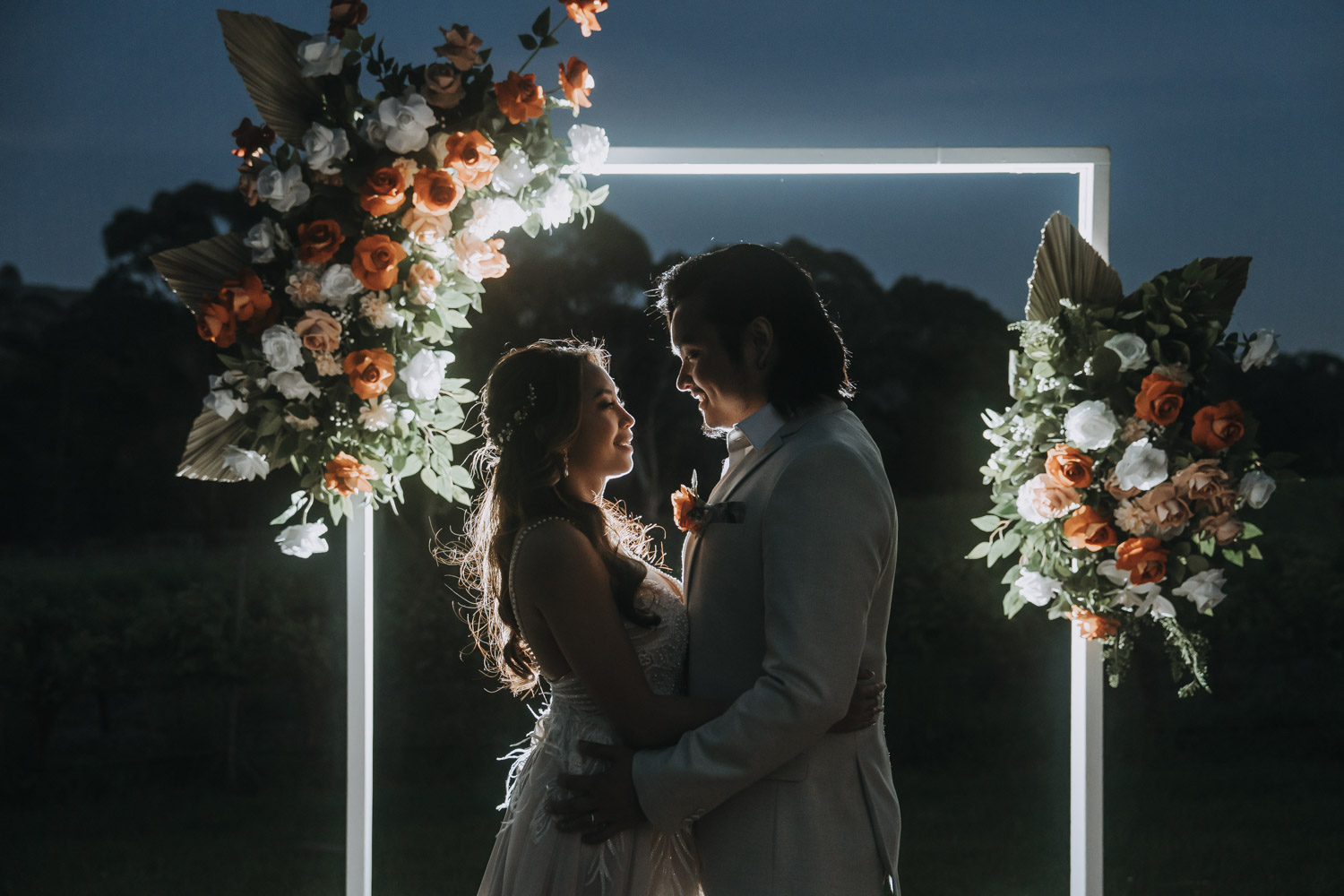 Bride and groom share a romantic moment under a floral archway with a backdrop of soft evening light, capturing the intimate ambiance of their wedding day by LaterStory. Melbourne wedding photography