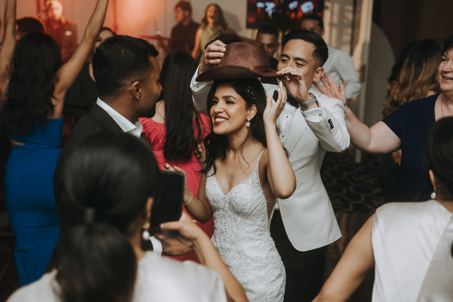 A Melbourne wedding photographer captures a joyous moment as the bride, donned in an exquisite lace gown, gleefully tries on a hat handed to her by a smiling guest. The groom, in a sharp white suit, looks on with affection amidst the bustling crowd of elegantly dressed guests, all immersed in celebration.