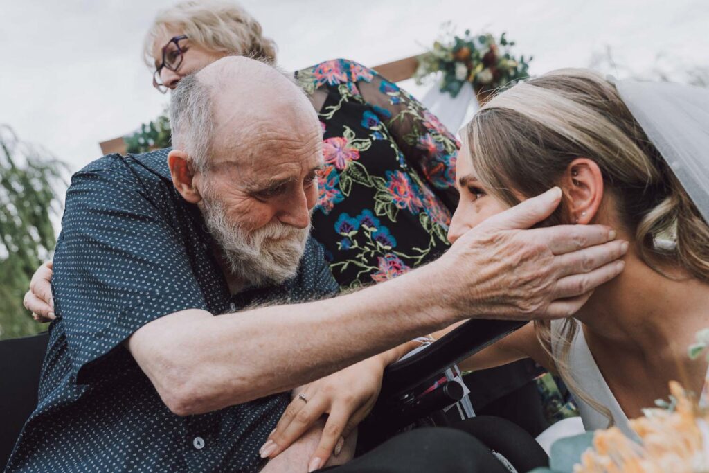 An emotive and tender moment captured in time: A bride, still in her wedding finery, leans forward to embrace an elderly gentleman seated in a wheelchair. His eyes are closed, and his face is a landscape of emotion as he holds her close. Behind them, a woman looks on, her expression a blend of happiness and nostalgic reflection. This powerful image, rich with storytelling, exemplifies the crucial and heartfelt shots that belong on any wedding photography checklist, ensuring that the quiet, yet profoundly meaningful moments are immortalized forever.