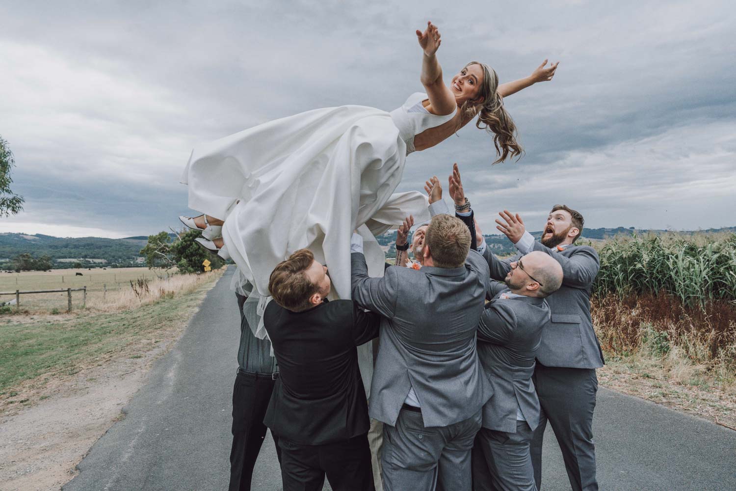 Bride joyfully being tossed into the air by groomsmen on a rural road near Melbourne, a lively moment captured during a wedding photoshoot in Melbourne.