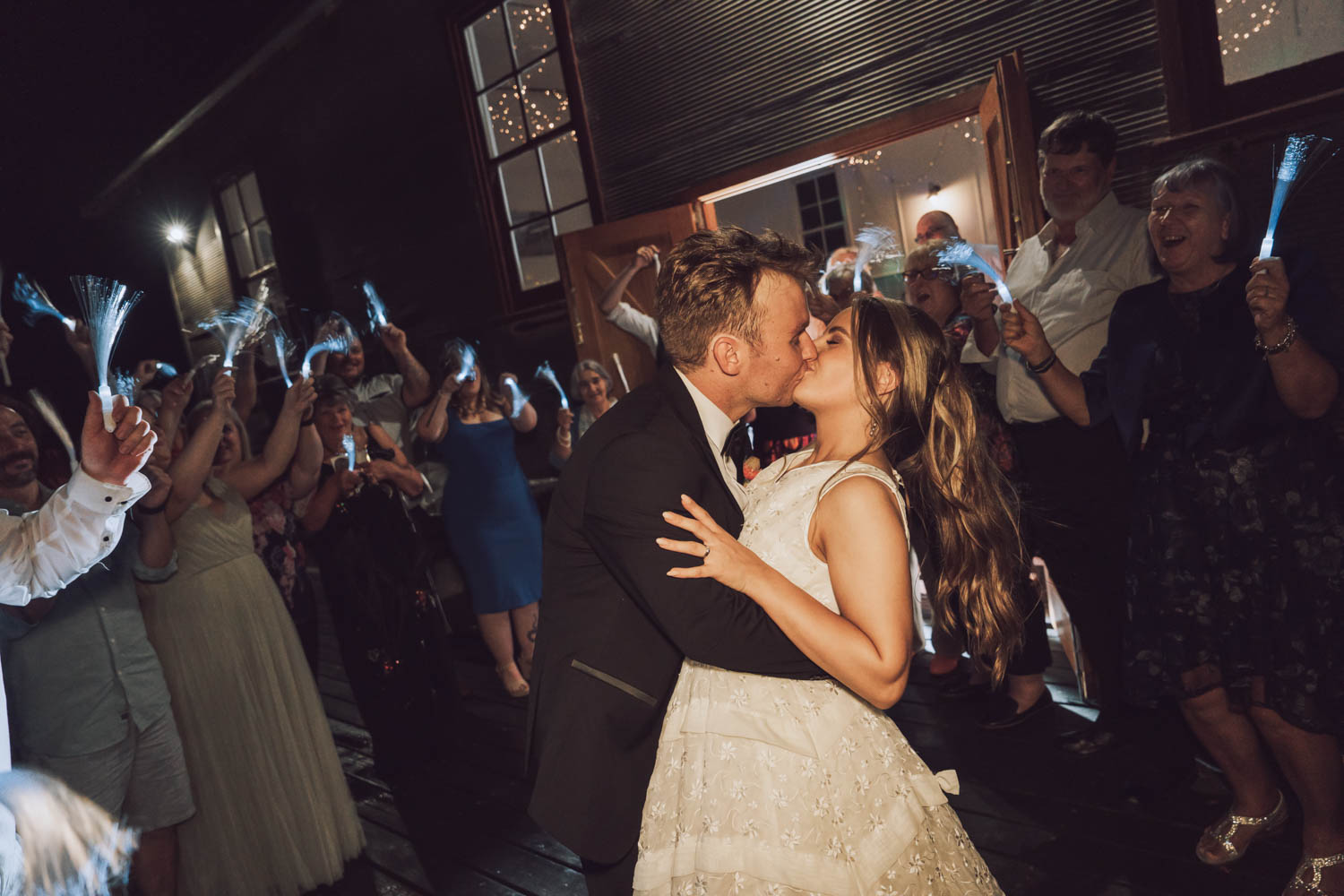 A heartfelt kiss between the bride and groom takes center stage against a backdrop of celebrating guests and sparkling lights, all immortalized by LaterStory affordable wedding photography.