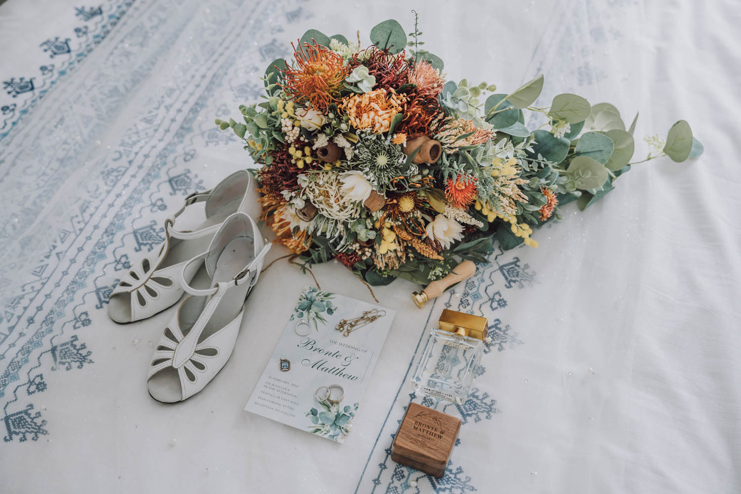 A beautifully arranged wedding photoshoot setup in Melbourne, featuring elegant bridal shoes, a vibrant bouquet, wedding invitations, and a perfume bottle, all laid out on a vintage patterned fabric.