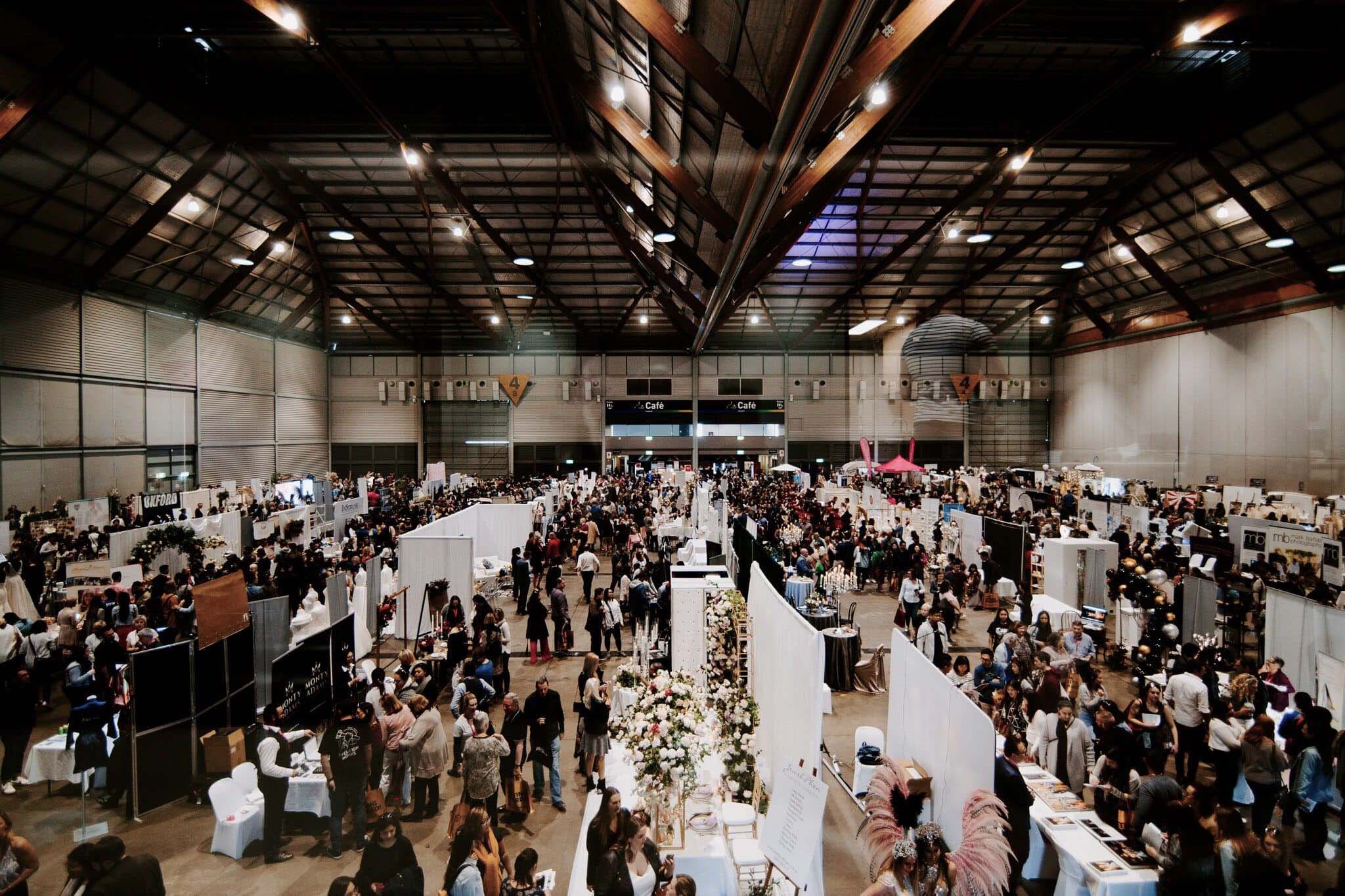 
A bustling indoor wedding expo with rows of vendor booths showcasing various services. Potential brides and grooms navigate through the crowd, exploring options for their special day. Prominently displayed are booths for wedding videography, capturing the attention of couples seeking to immortalize their wedding memories.