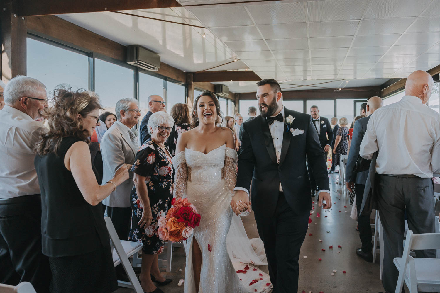 A beaming bride and groom hold hands as they joyfully walk down the aisle post-ceremony, encapsulating the perfect moment for a wedding photographer to capture. The bride, in a glittering off-shoulder gown, and the groom, in a classic black tuxedo, exude happiness amidst a shower of rose petals. Guests line the aisle, some clapping, others wiping away tears, sharing in the couple’s happiness. The warmth and love of the occasion are palpable, with every detail from the guests' attire to the bright bouquets enhancing the celebratory mood. This image is a quintessential example of the candid, heartfelt shots that a skilled wedding photographer aims to capture, preserving the genuine emotions of a wedding day.