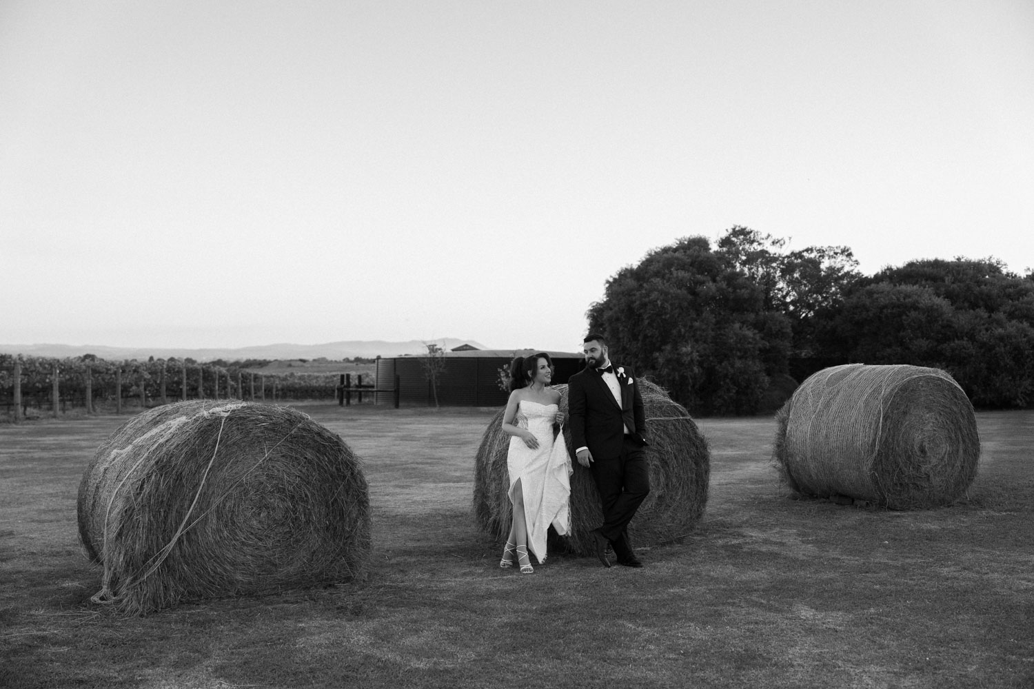 A Melbourne wedding photographer captures a timeless black and white image of a newlywed couple walking hand in hand beside large hay bales in a rustic field, symbolizing the start of their journey together, with the tranquil expanse of the Australian countryside stretching out around them.