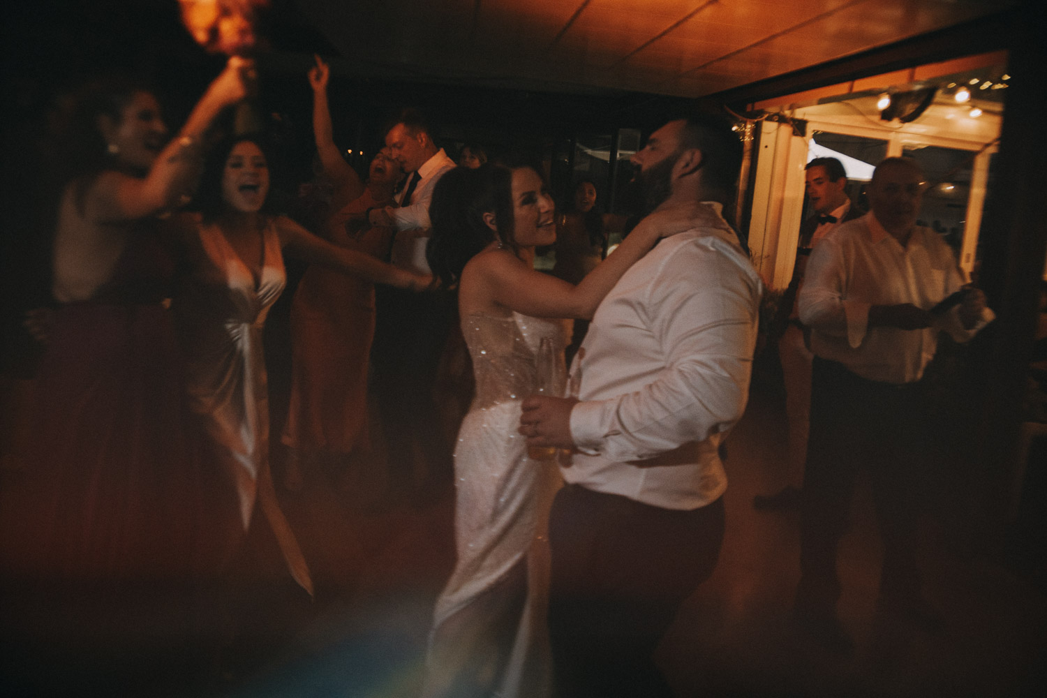 In the soft, warm glow of the celebration, a bride in a shimmering gown dances closely with her groom, whose shirt echoes the intimacy of the moment with its sleeves casually rolled up. They are surrounded by jubilant guests, lost in the revelry of dance, their joyous expressions and lively movements creating an atmosphere of unbridled festivity. This candid capture by a wedding photographer showcases the vivacious spirit of a reception, a testament to the photographer's ability to encapsulate the essence of the couple’s joyous day through spontaneous and emotive imagery.