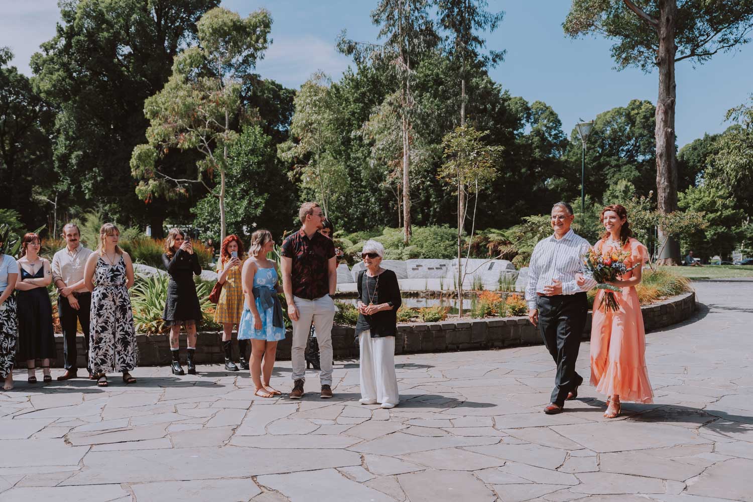 A budget wedding ceremony held outdoors with a bride in a flowing peach dress walking alongside an older man, surrounded by guests in casual and colorful attire, all gathered on a stone-paved area with lush greenery in the background.