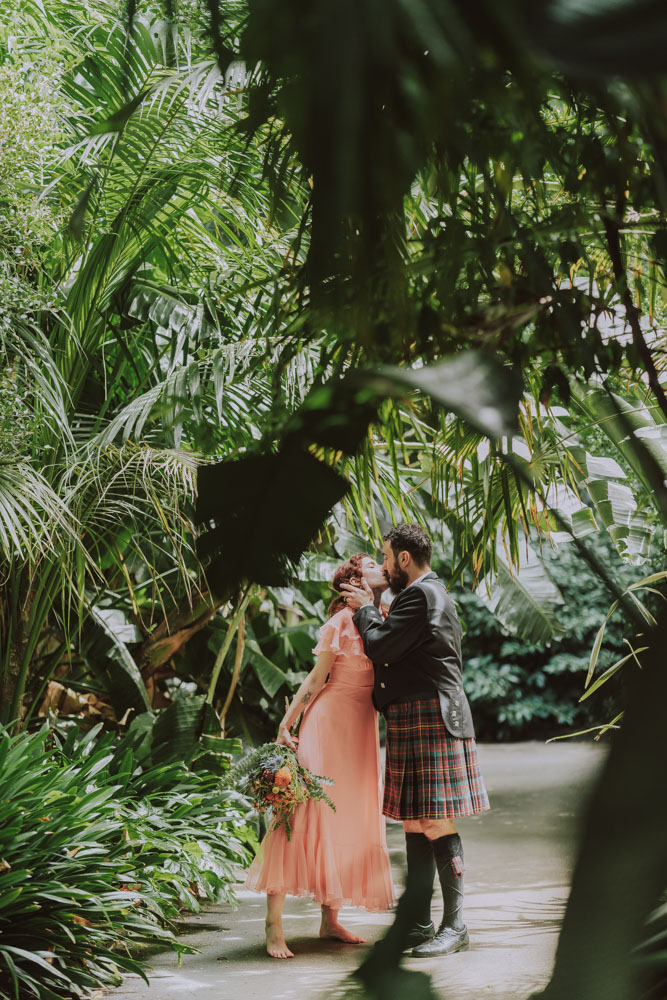 A tender moment at a budget wedding captured in a lush, tropical garden setting, featuring a bride in a long peach dress and a groom in a tartan kilt, sharing a kiss surrounded by dense green foliage.