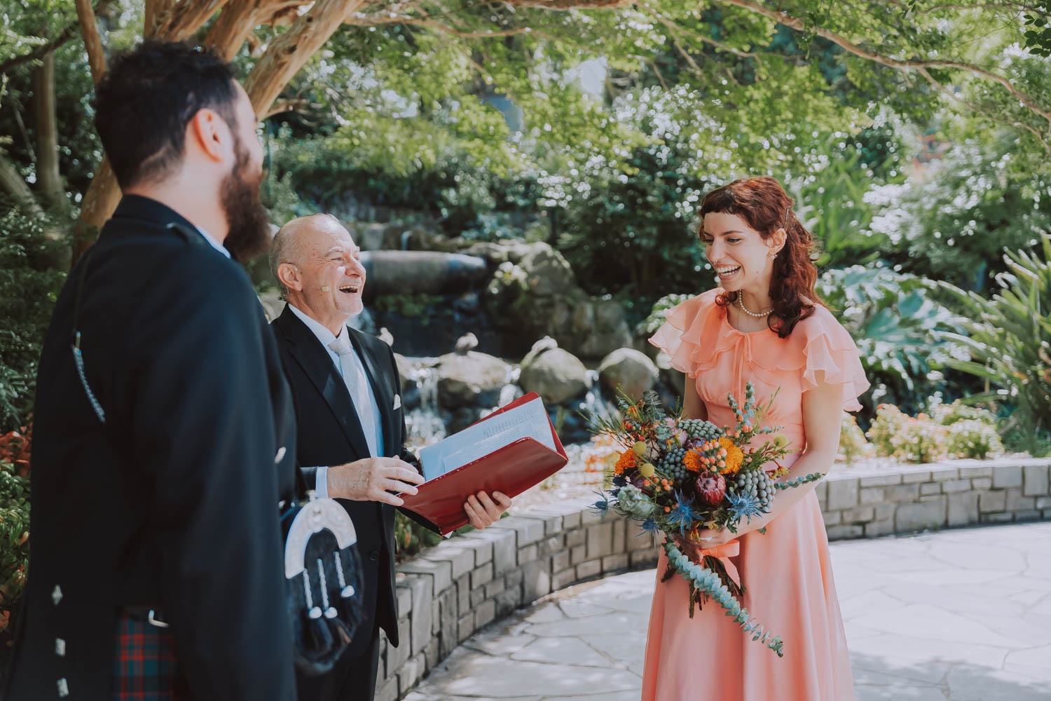 A joyful moment at a budget wedding ceremony with the bride in a peach dress laughing beside a celebrant holding a red book, and a groom in a kilt, all standing in a lush garden setting with a waterfall in the background.